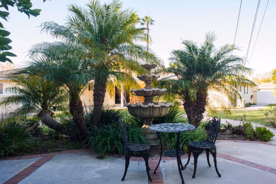 Outdoor patio with a three-tier fountain, palm trees, and a metal bistro table with two chairs in front of a single-story building.