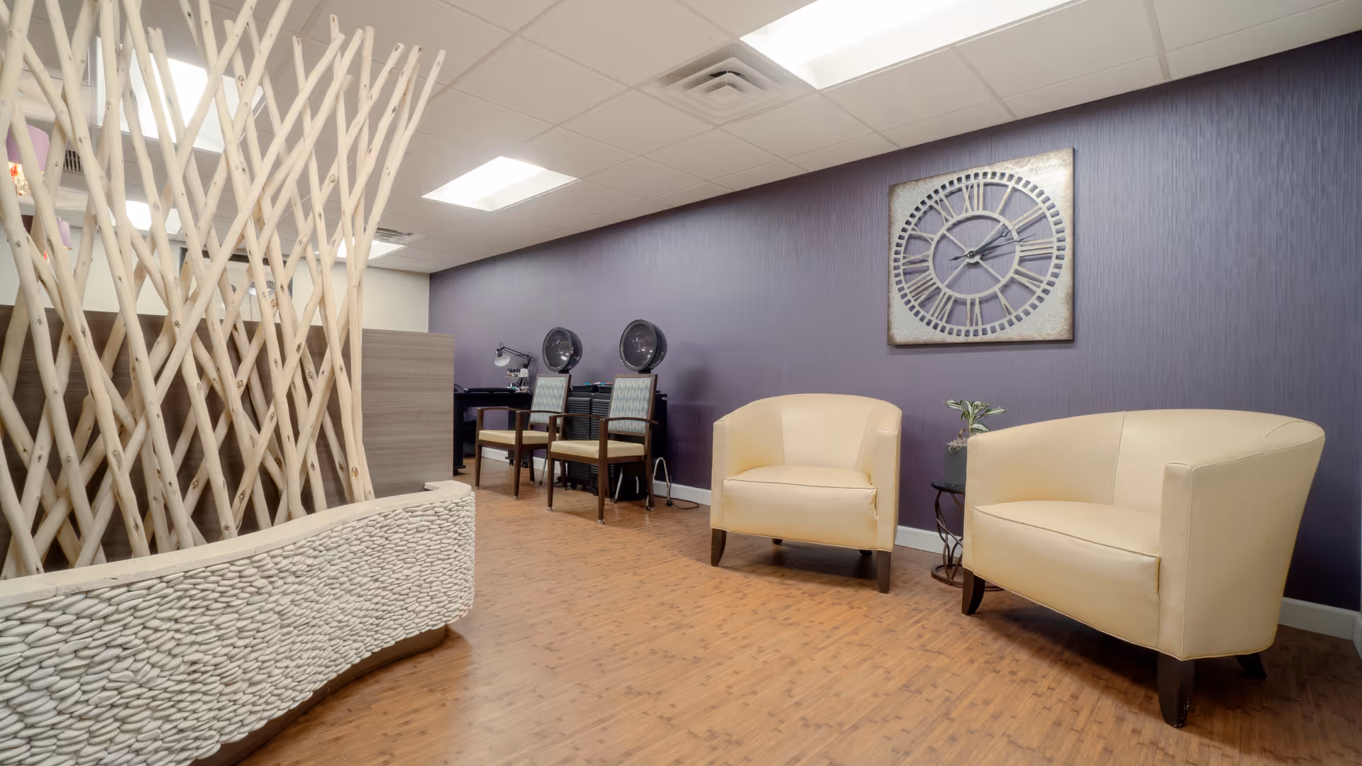 A modern waiting area with two beige armchairs and a small round table with a plant between them, set against a purple wall with a large decorative clock. The floor is wooden, and there are two chairs with hair dryers in the background, separated by a decorative wooden partition.