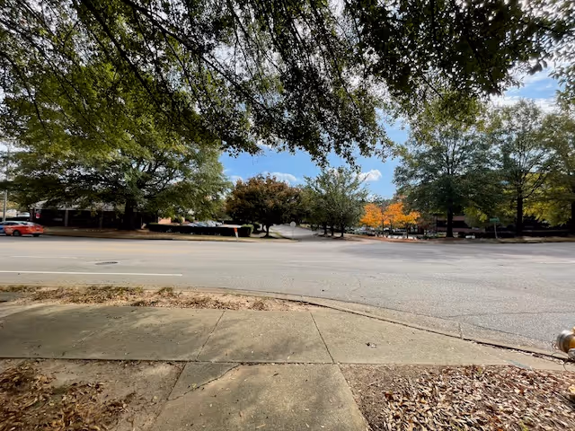 View across a quiet street toward a tree-lined parking area and entrance with autumn foliage under a partly cloudy sky.