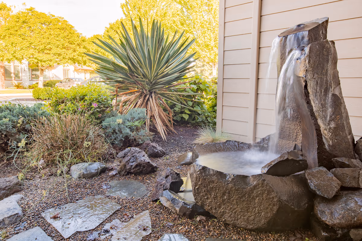 Stone water fountain by a building with a rock garden and plants.