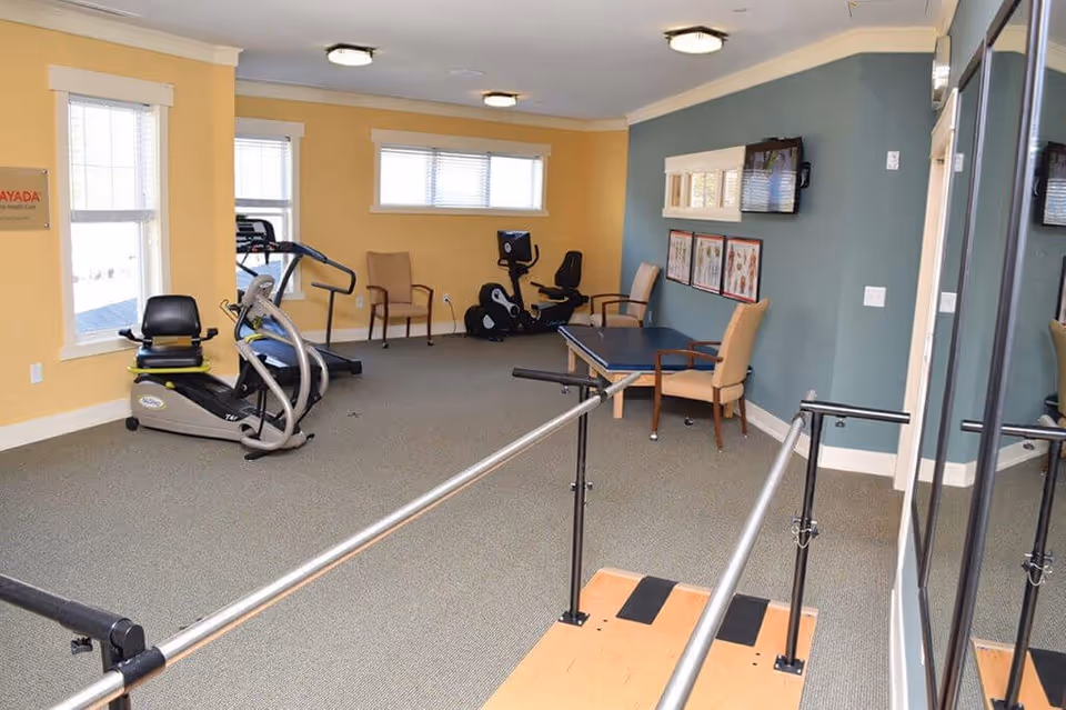 A bright therapy/exercise room with stationary exercise machines, parallel bars, chairs, and a wall-mounted TV.