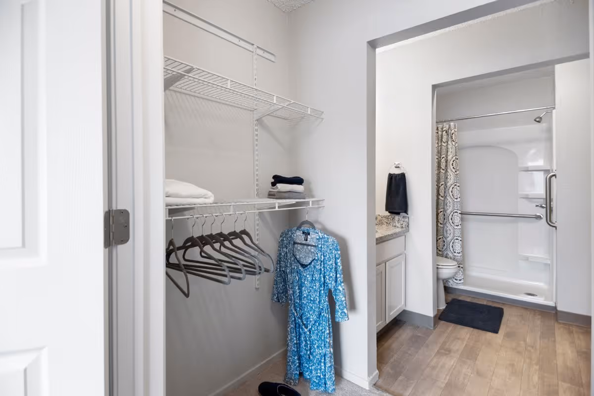 View of a closet with wire shelving holding folded towels and empty hangers, next to a bathroom with a shower, toilet, and sink with granite countertop. A blue patterned dress hangs on one of the hangers in the closet.
