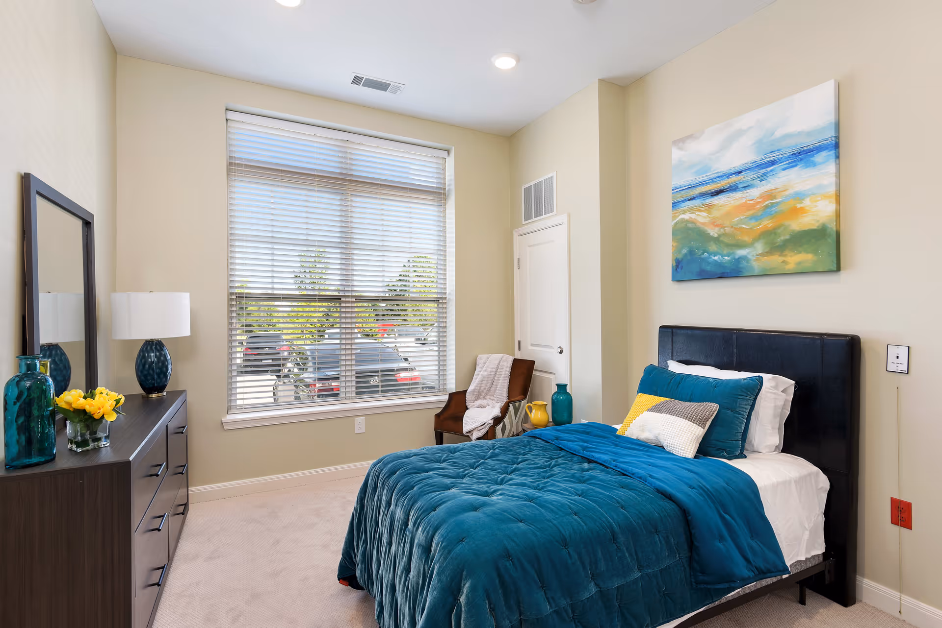 A bedroom with a large window covered by blinds, a bed with teal bedding and multiple pillows, a dark wood dresser with a mirror, a lamp, and decorative vases. There is a chair with a blanket and a colorful abstract painting on the wall above the bed.