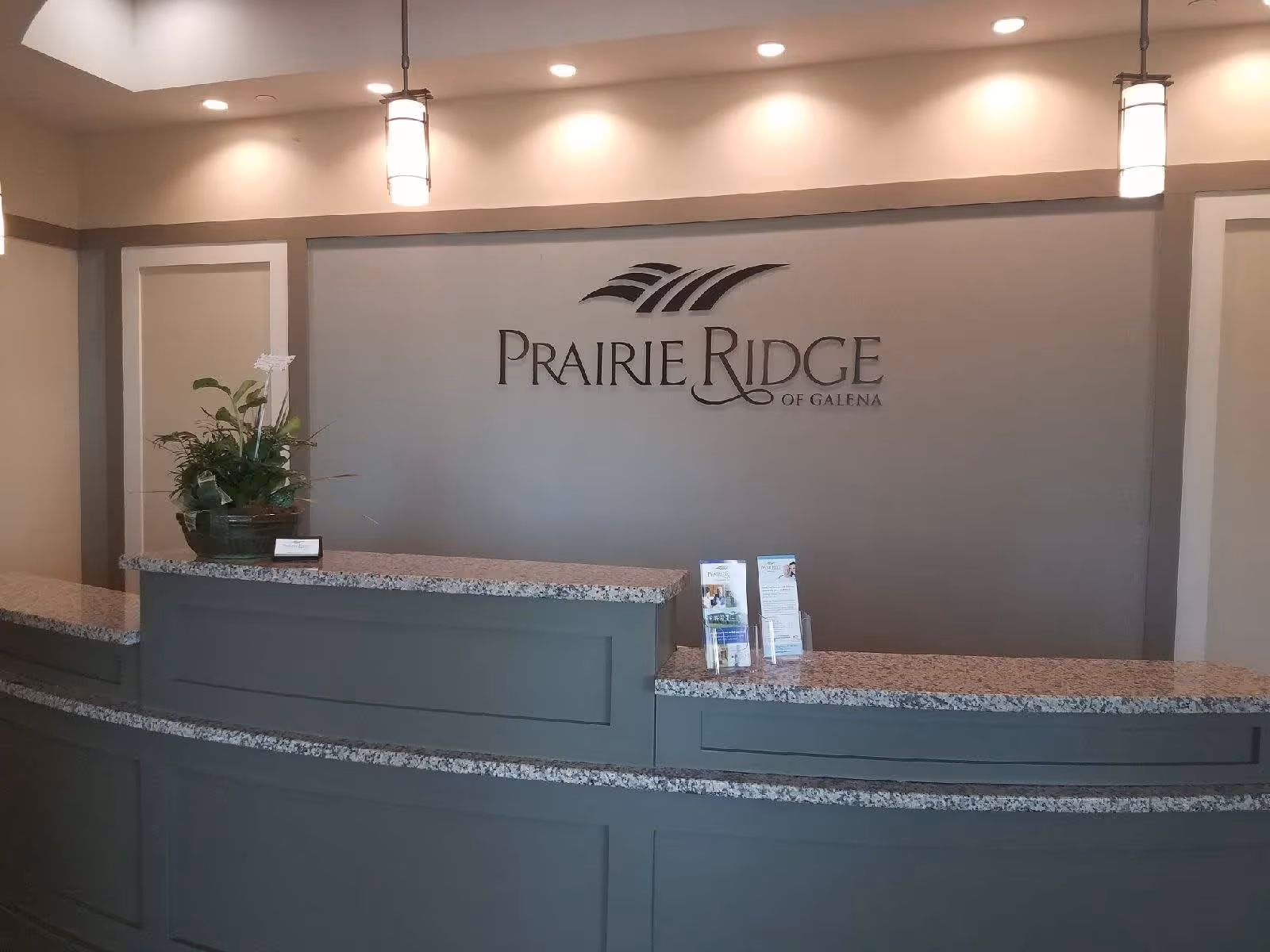 Reception desk and logo wall of Prairie Ridge of Galena with granite countertop, pendant lights, a potted plant, and brochures.
