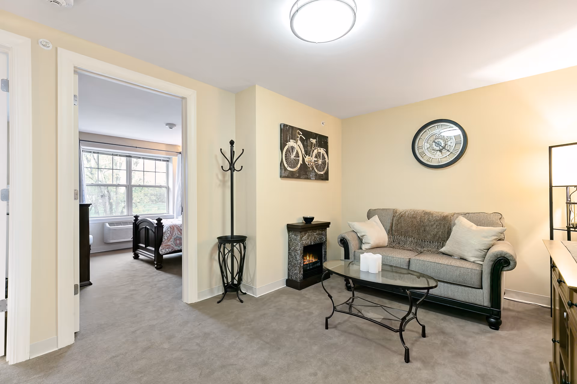 A cozy living room with a gray sofa adorned with two pillows and a throw blanket, a glass coffee table with three white candles, a small electric fireplace, a coat rack, and wall decorations including a bicycle painting and a round clock. An adjacent bedroom with a large window and a bed is visible through an open doorway.