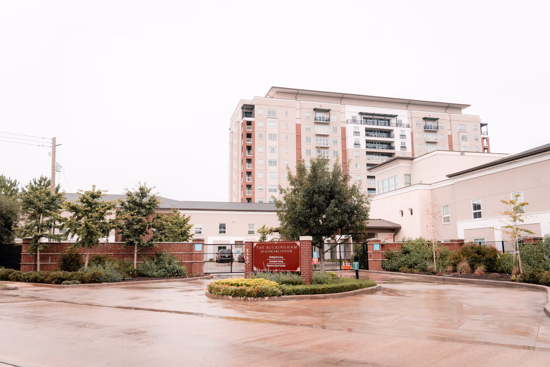 Exterior view of The Buckingham Healthcare Center showing a multi-story building behind a gated entrance with a red brick sign displaying the facility name and services including Skilled Nursing, Assisted Living, and Memory Care Center. The area in front is a wet driveway with some landscaping and trees.