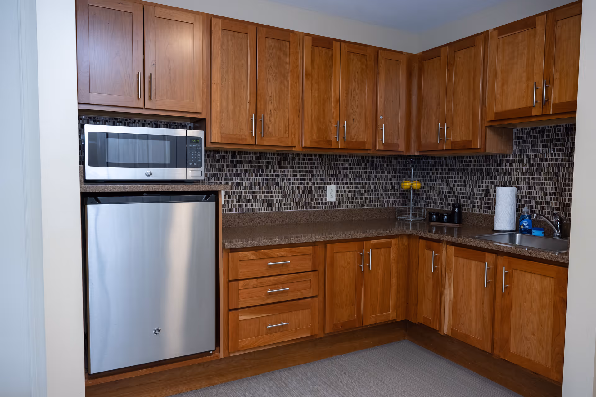 A kitchen area with wooden cabinets, a stainless steel microwave and mini refrigerator, a brown countertop, a small sink with a faucet, paper towels, dish soap, and a tiled backsplash in shades of brown and gray.