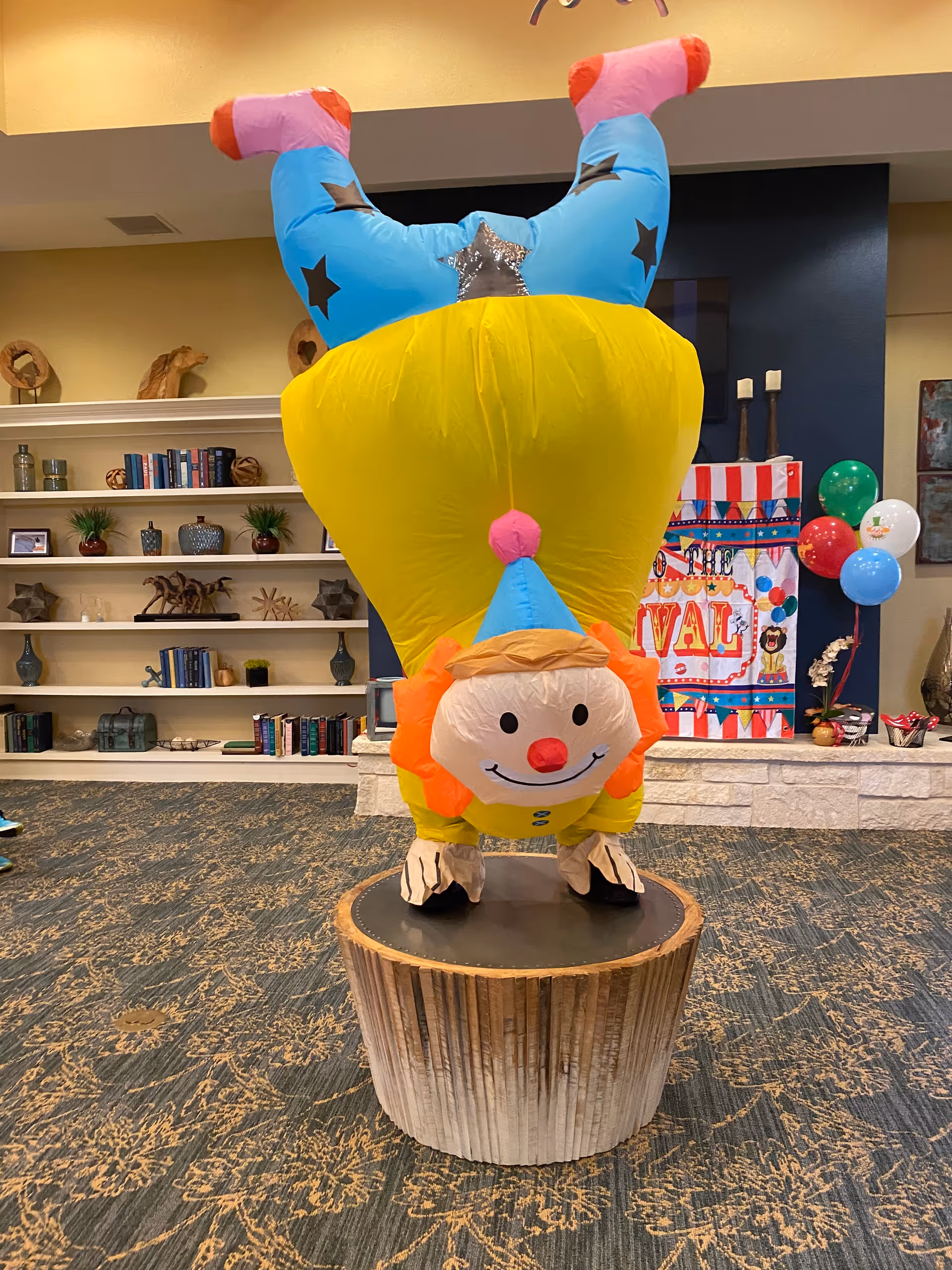 A large upside-down inflatable clown standing on a wooden stump pedestal in a decorated community room with bookshelves and balloons.