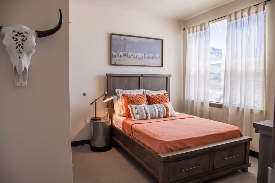 A cozy bedroom with a wooden bed frame and coral-colored bedding, accented with multiple pillows. A bedside table with a lamp and a glass of water is next to the bed. A decorative skull with horns is mounted on the wall to the left, and a framed picture of sheep hangs above the bed. Light filters through sheer curtains covering a window on the right.