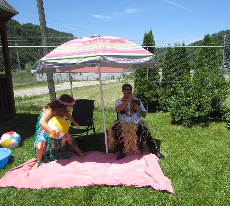 Two people dressed in tropical-themed attire sitting and standing on a pink blanket under a striped umbrella in a grassy outdoor area. One person is sitting on a chair wearing sunglasses and a grass skirt, holding a pineapple cup, while the other person is standing holding a beach ball. There is a small blue kiddie pool and another beach ball nearby, with a chain-link fence and trees in the background.