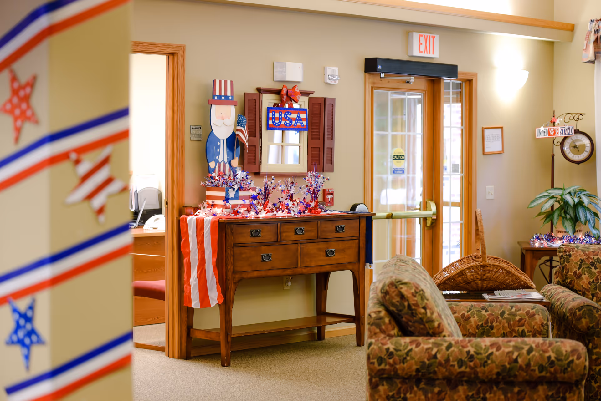 A cozy interior space decorated with patriotic American-themed decorations including a wooden Uncle Sam figure, a USA sign, and red, white, and blue garlands. There is a wooden table with drawers, a floral patterned armchair, a wicker basket, and a clock on a stand. A glass door with an exit sign is visible in the background.