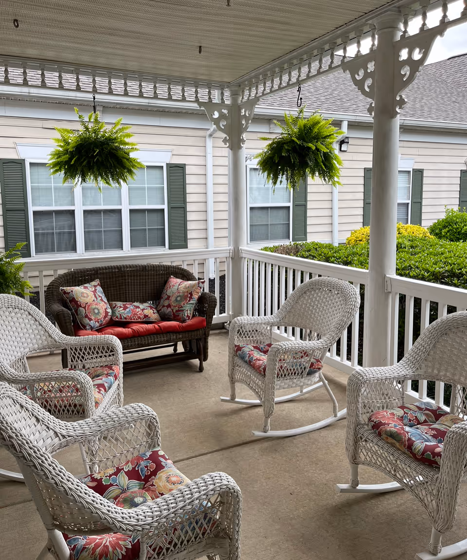 Covered outdoor porch area with white wicker rocking chairs and a dark wicker loveseat, all with floral cushions. Two hanging green ferns are suspended from the porch ceiling. The porch overlooks a garden with bushes and a beige building with green shutters and windows in the background.