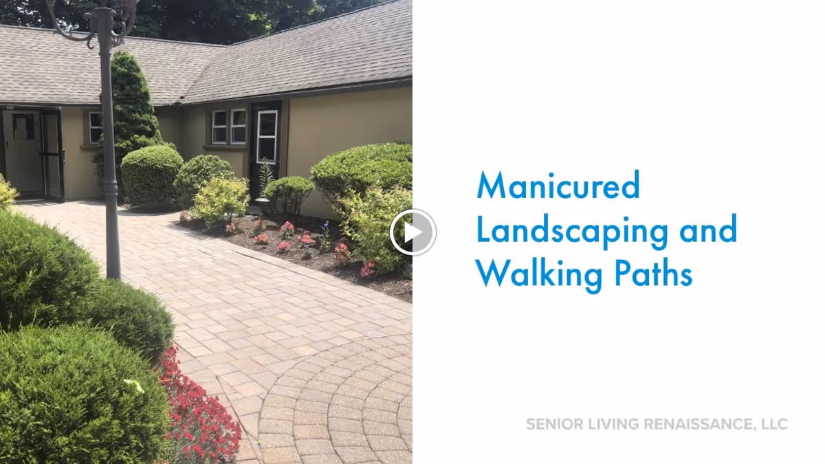 Outdoor paved walking path with manicured bushes and flowers alongside a single-story building with beige walls and multiple windows under a shingled roof.