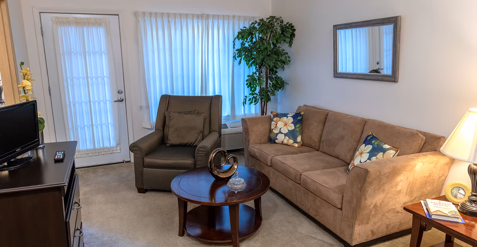 A cozy living room with a beige sofa adorned with floral pillows, a dark armchair with a matching cushion, a round wooden coffee table with decorative items, a TV on a dark wooden stand, a side table with a lamp and books, a large window with white curtains, and a potted plant in the corner.