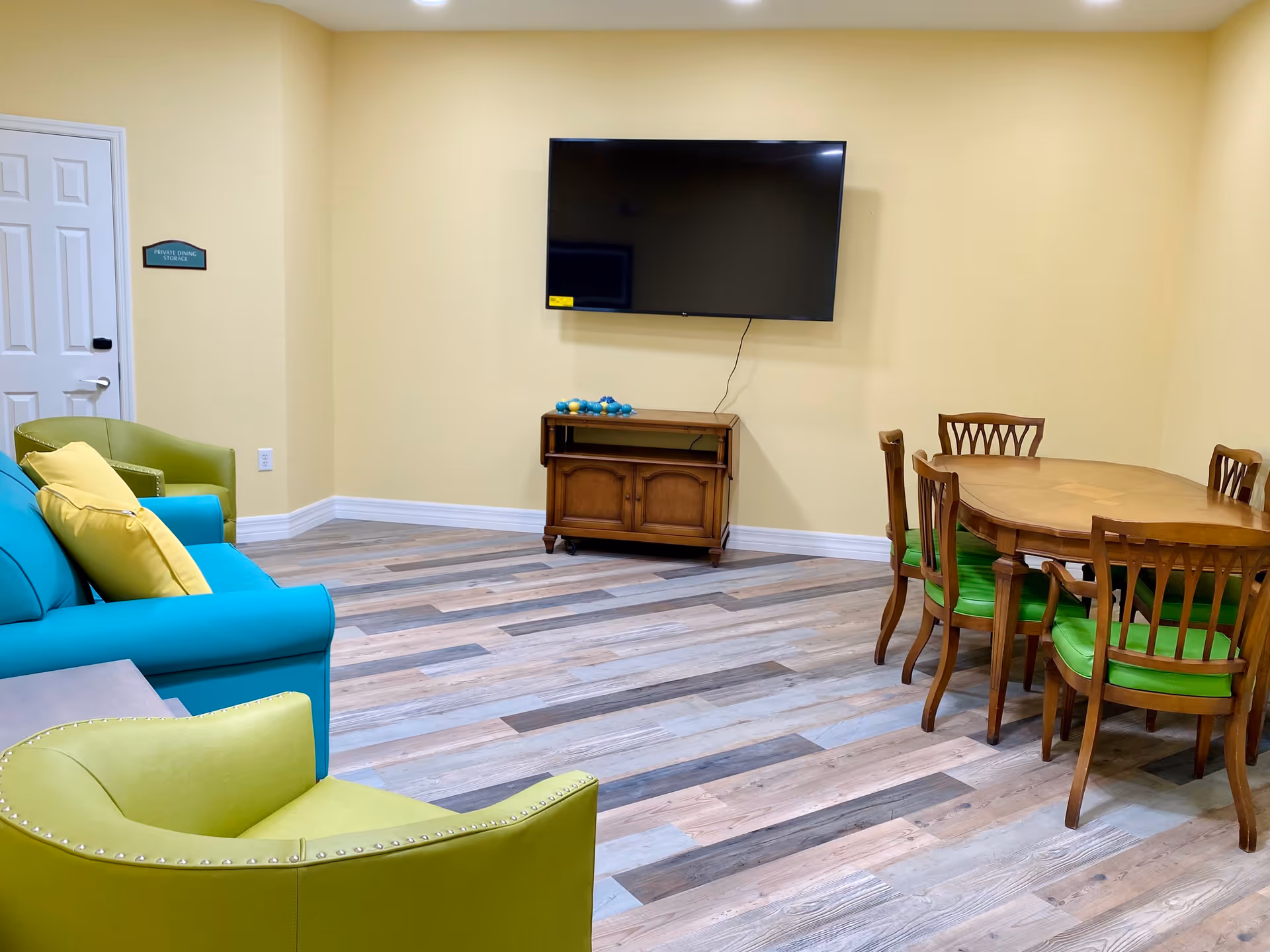 A room with light yellow walls and wood-patterned flooring featuring a turquoise sofa with yellow pillows, two green armchairs, a wooden dining table with six chairs that have green cushions, a wall-mounted flat-screen TV above a small wooden cabinet, and a door labeled 'Private Dining Storage'.