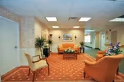 Carpeted lounge area with upholstered chairs and a sofa arranged around a coffee table, potted plants, and framed art on the wall.