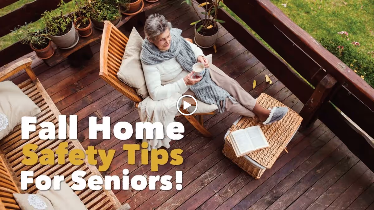 An elderly woman sitting comfortably on a wooden chair on a porch or deck, wrapped in a cozy scarf and holding a cup. There are several potted plants on a wooden shelf nearby, and a wicker table with an open book and slippers resting on it. The text overlay reads 'Fall Home Safety Tips For Seniors!'