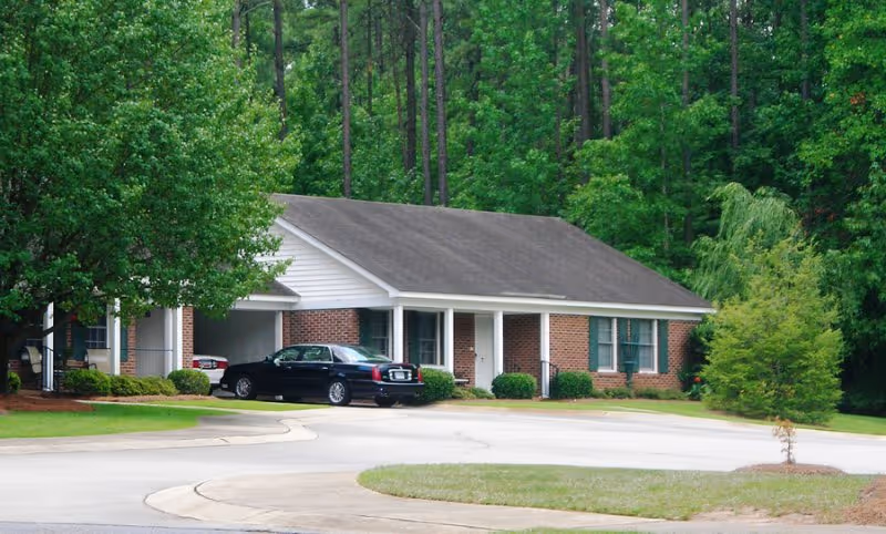 Single-story brick building with a covered carport and a black sedan parked in front, set among green trees and lawn.