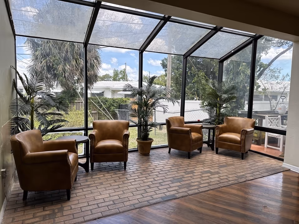 A sunroom with large glass windows and ceiling panels overlooking a garden area. Inside, there are four brown leather armchairs arranged around two small black tables. Several potted green plants are placed near the windows, and the floor is a combination of brick and wood.