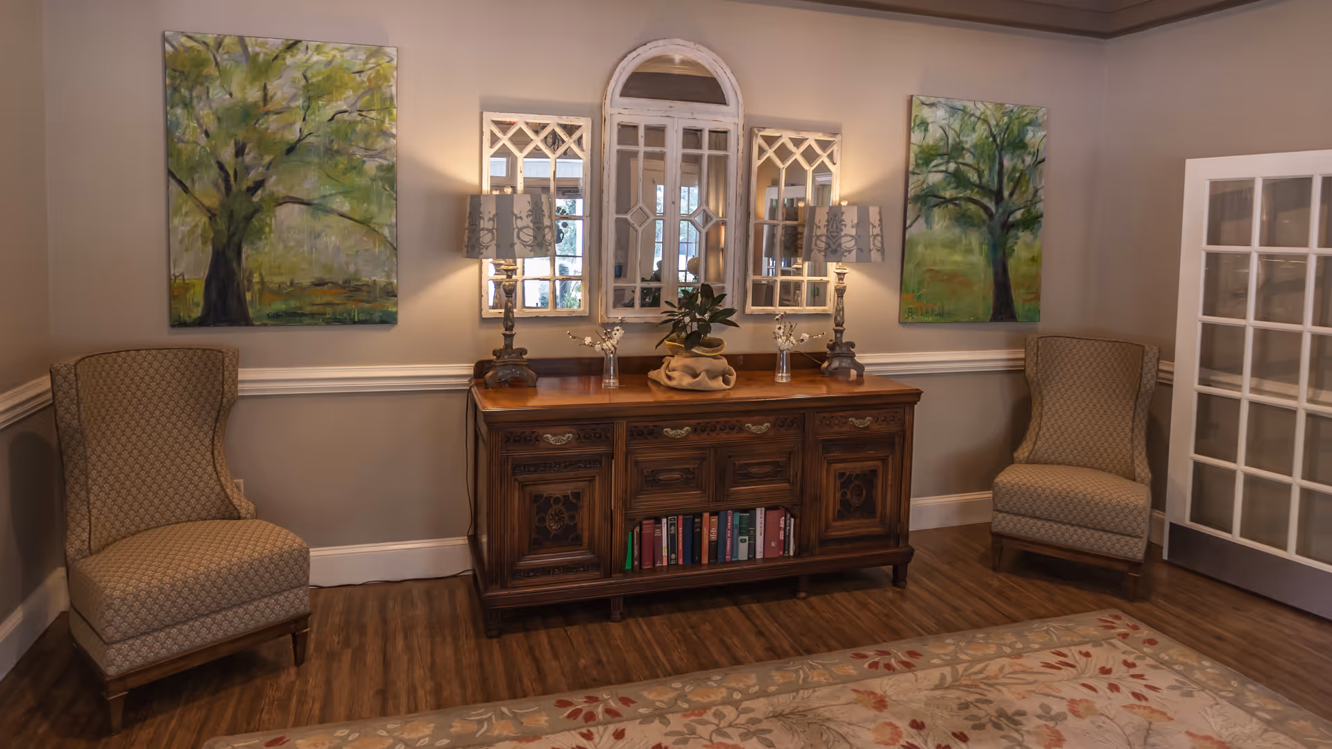 A cozy interior corner of a living space featuring two patterned armchairs on either side of a wooden sideboard with books on the lower shelf. On top of the sideboard are two decorative lamps, a small plant, and two glass vases. Above the sideboard are three decorative mirrors with windowpane designs. The walls are adorned with two paintings of green trees. The floor is wooden with a floral area rug partially visible, and a white-framed glass door is on the right side.