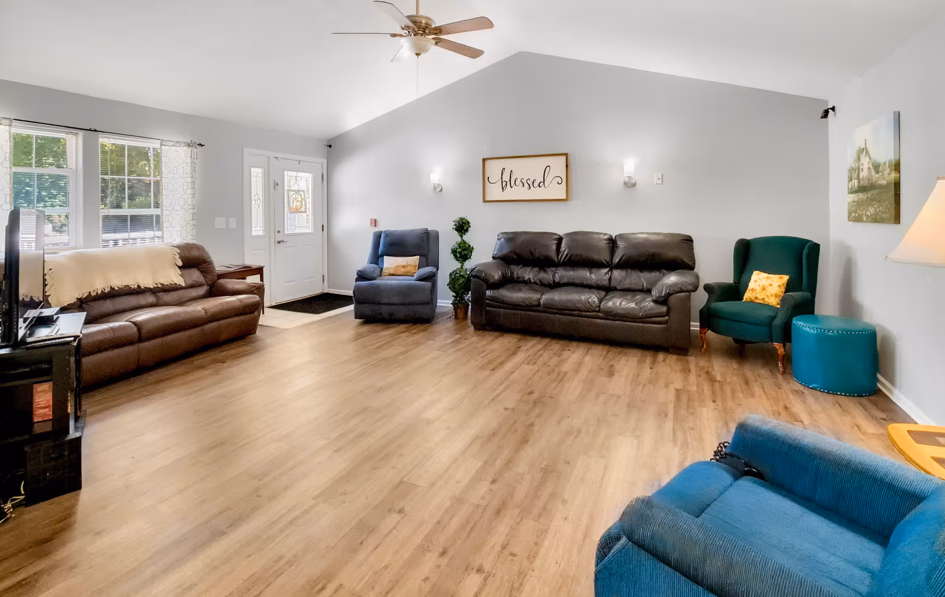 A spacious living room with wooden flooring, featuring a brown leather sofa, a blue recliner chair, a dark brown leather couch, a green armchair with a yellow pillow, and a teal ottoman. The room has a ceiling fan, a wall-mounted sign that says 'blessed', a lamp, and a TV on a stand near a window with white curtains. The front door with glass panels is visible in the background.