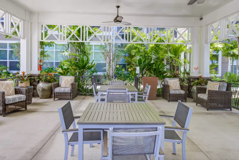 Covered outdoor seating area with tables, chairs, and wicker lounge chairs surrounded by tropical plants.