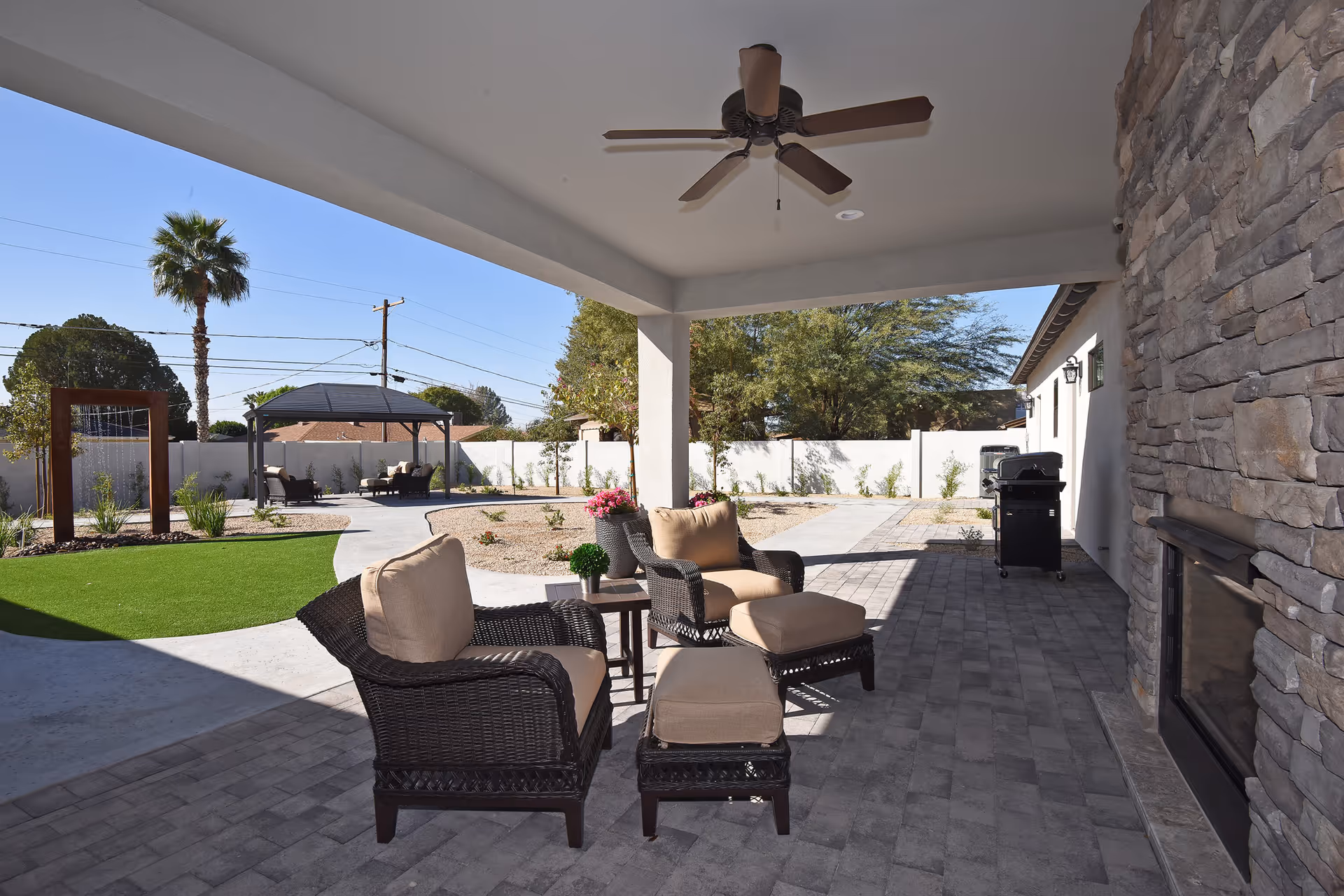 Covered outdoor patio area with cushioned wicker chairs, ottomans, a small table, and a ceiling fan. The patio overlooks a landscaped yard with a gazebo, palm trees, and a water feature, all enclosed by a white fence.