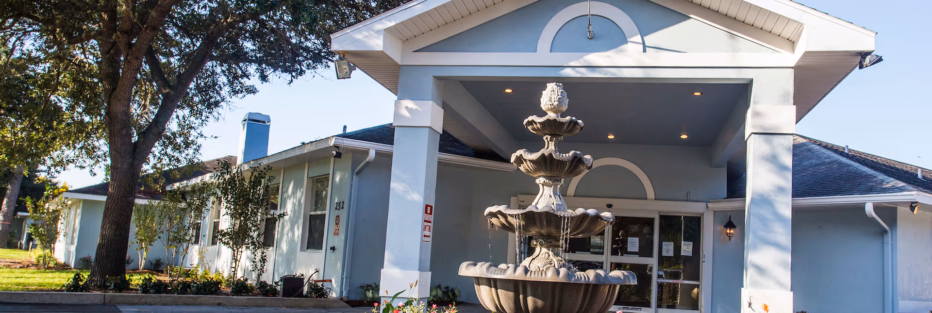 Exterior view of Magnolia Manor senior living facility entrance with a multi-tiered water fountain in front, surrounded by trees and landscaping under a clear sky.