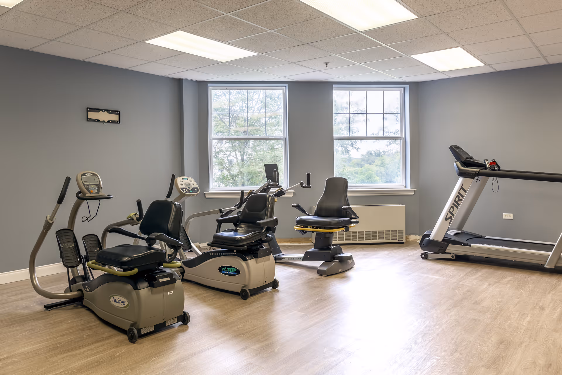 A fitness room with exercise equipment including two recumbent stationary bikes, an elliptical machine, and a treadmill. The room has light wood flooring, gray walls, a white ceiling with fluorescent lights, and two large windows showing trees outside.