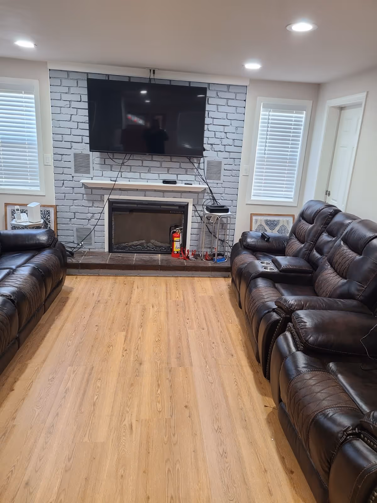 A living room with two dark brown leather reclining sofas facing each other across a wooden floor. A flat-screen TV is mounted on a white brick wall above a fireplace. There are two windows with white blinds on either side of the fireplace, and a fire extinguisher is placed on the hearth.