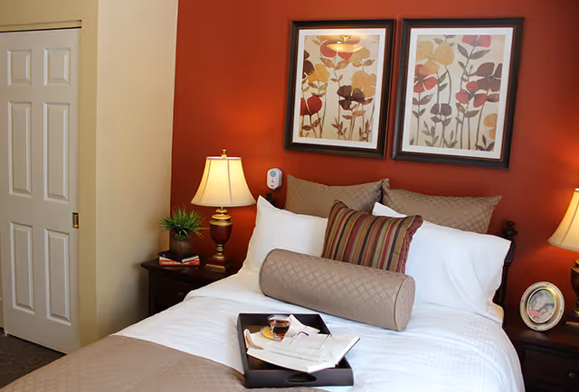 A neatly made bed with white sheets, beige and striped pillows, and a beige quilted bolster pillow in a bedroom with a red accent wall. Two framed floral artworks hang above the bed. There are two bedside tables with lamps, a small plant, books, and a framed photo. A tray with a cup of tea, an open book, and reading glasses rests on the bed.