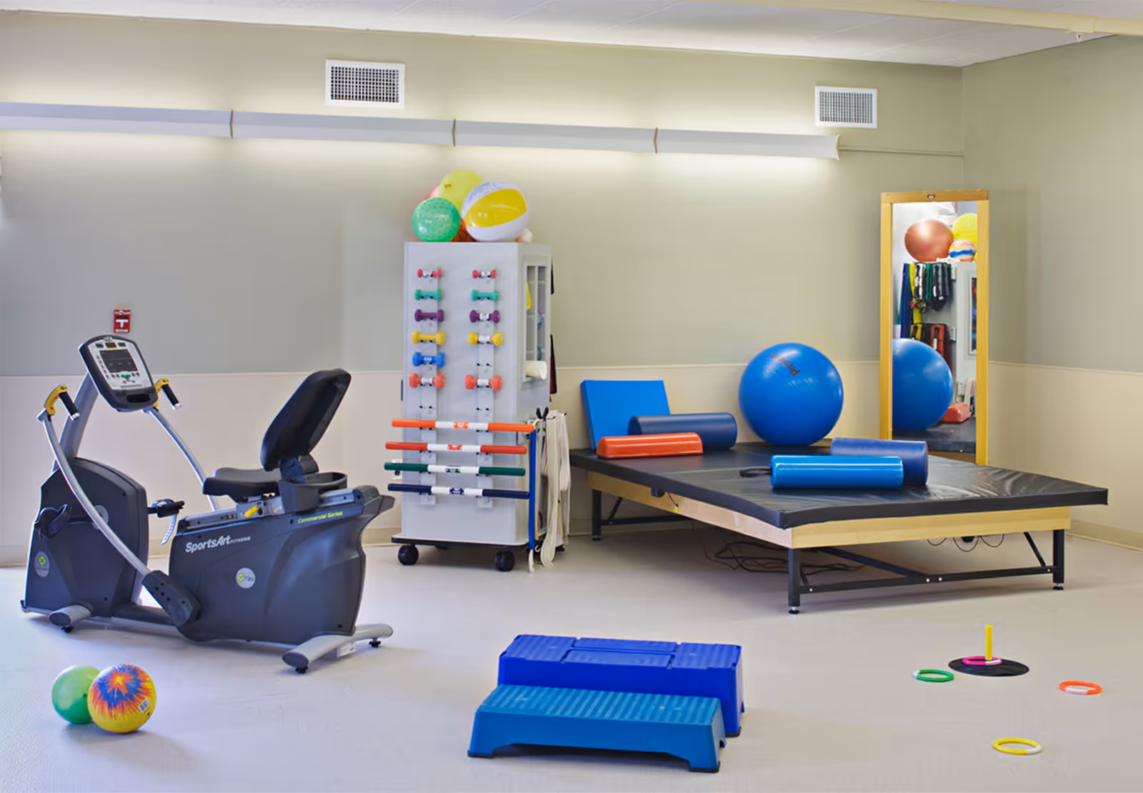 Therapy/exercise room with a recumbent bike, treatment table, exercise balls, small weights, and step platforms.