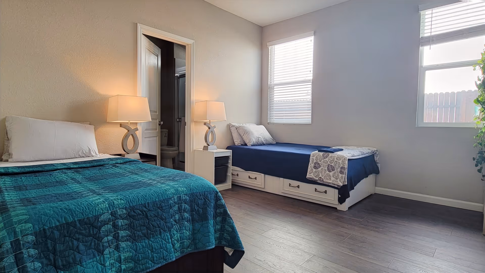 A bedroom with two single beds, one with a teal quilt and the other with a navy blue quilt. Each bed has pillows and a bedside table with a lamp. There is a window with blinds letting in natural light, and a door leading to a bathroom with a visible toilet. The floor is wooden, and there is a plant near the window.
