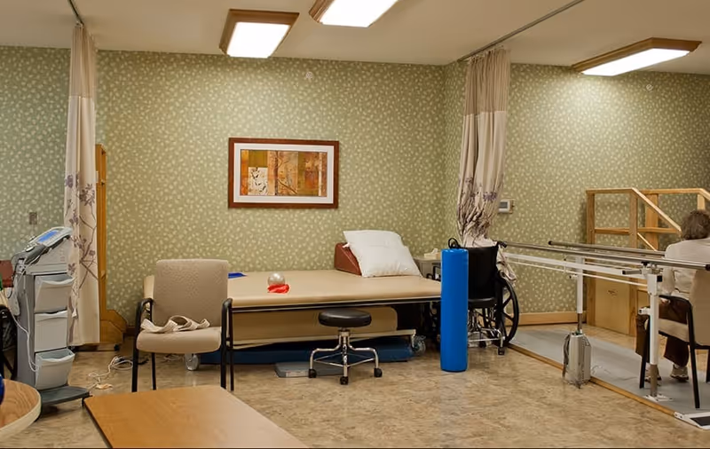 Rehabilitation room with a treatment table, wheelchair, parallel bars, chairs, and therapy equipment against patterned wallpaper.