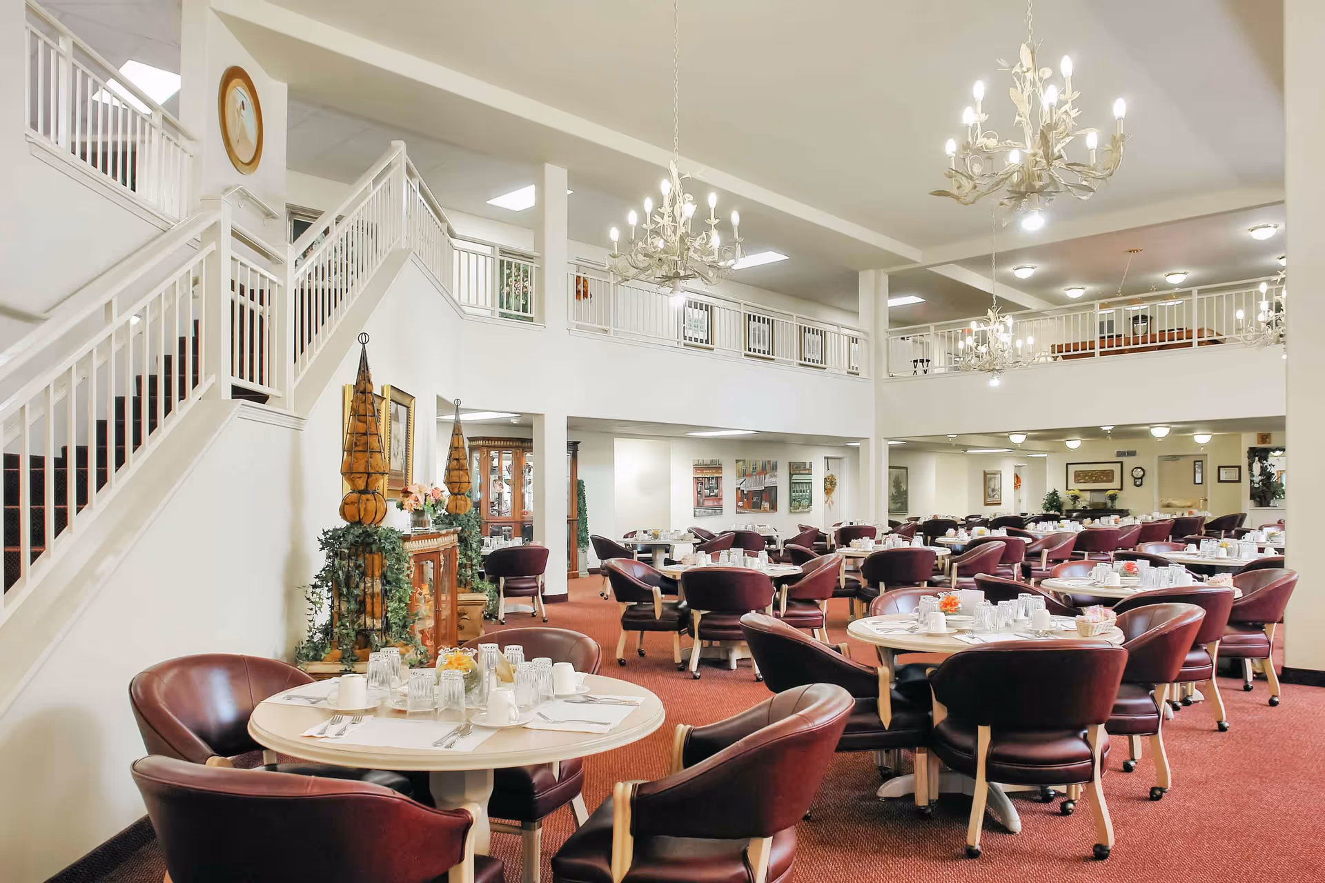 Spacious two-story dining room with round tables set for meals, burgundy chairs, chandeliers, and a staircase leading to an upper balcony.