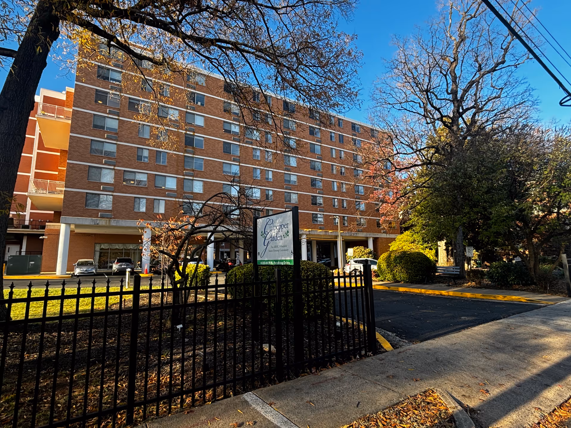 Exterior view of a multi-story brick building with numerous windows, identified as Culpepper Garden III. The building is surrounded by trees with autumn foliage, a black metal fence, and a sidewalk. A sign in front of the building displays the name Culpepper Garden.