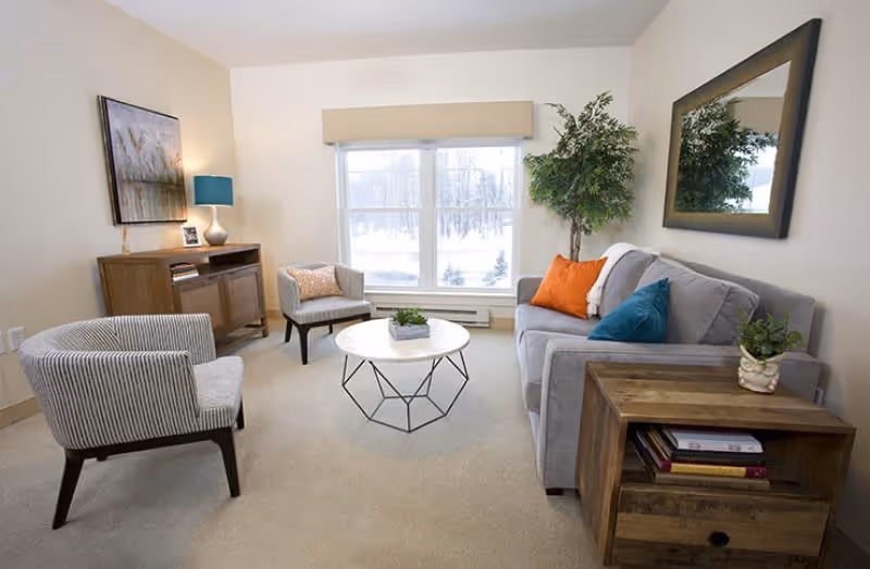 A cozy living room with a gray sofa adorned with orange and blue pillows, two striped armchairs, a round coffee table with a small plant, a wooden side table with books and a plant, a wooden cabinet with a lamp and framed picture, a large mirror on the wall, and a window showing a snowy outdoor scene.