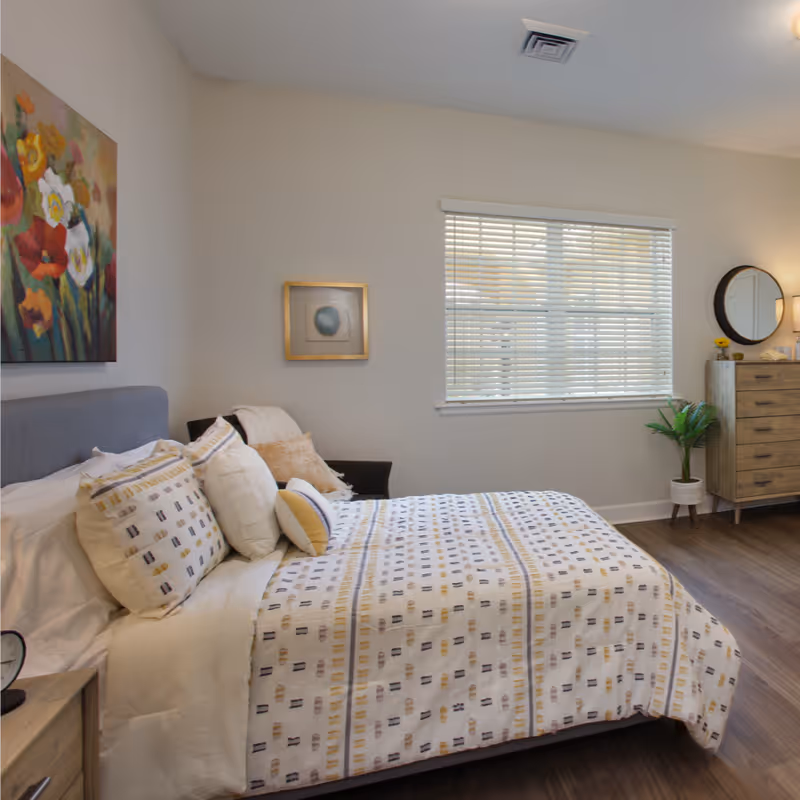 A neatly made bed with patterned bedding and multiple pillows in a bright bedroom. The room features a window with blinds, a wooden dresser with a round mirror above it, a small potted plant, and colorful floral artwork on the wall.
