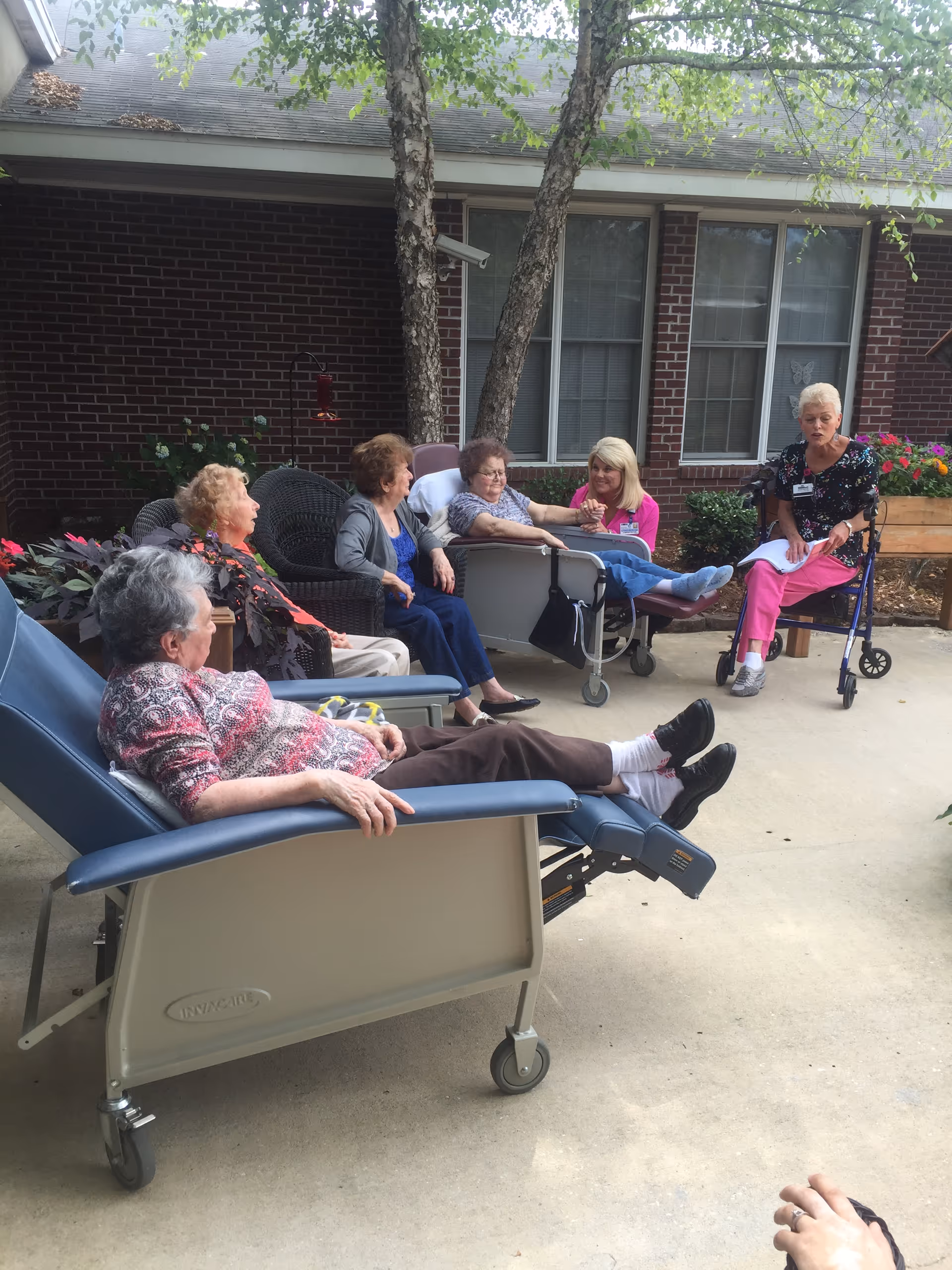 Several elderly residents and a caregiver sitting and reclining in a nursing home courtyard in front of a brick building.