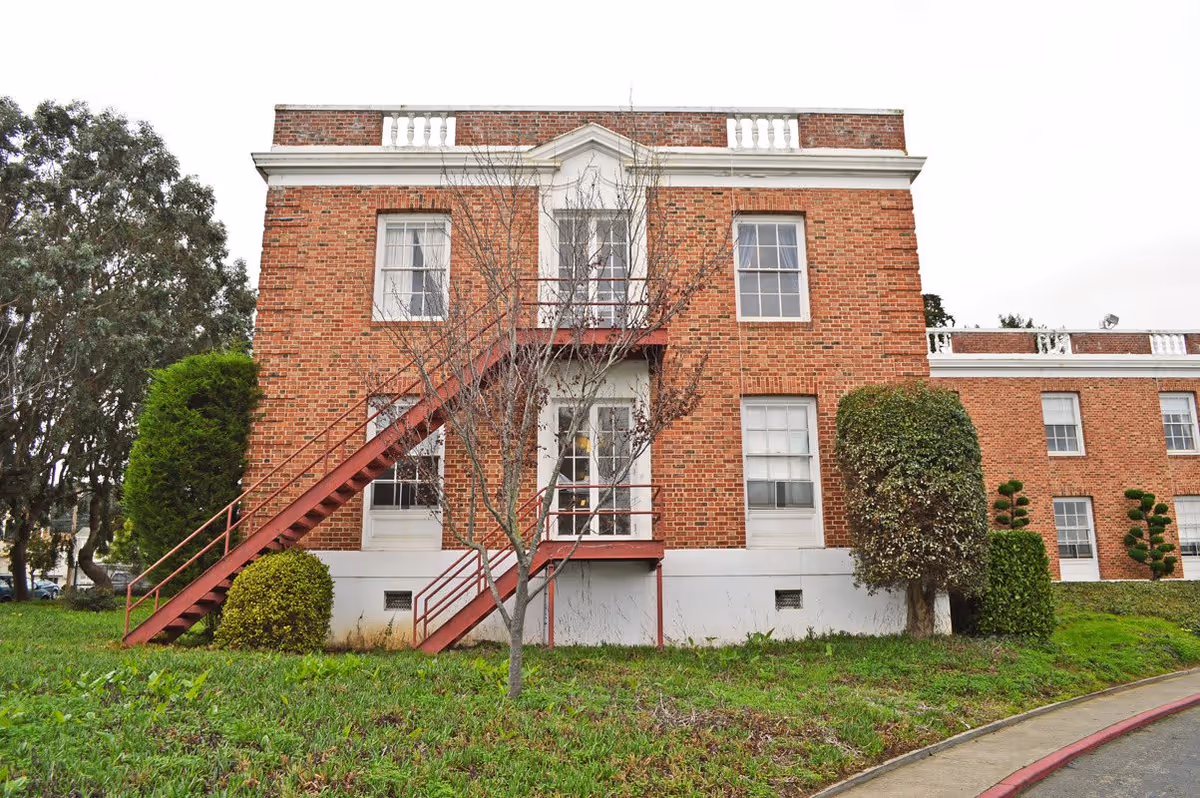 Exterior view of a two-story brick building with white trim and multiple windows. There is a red metal staircase leading to a door on the second floor. The building is surrounded by green grass, trimmed bushes, and a leafless tree in the foreground.