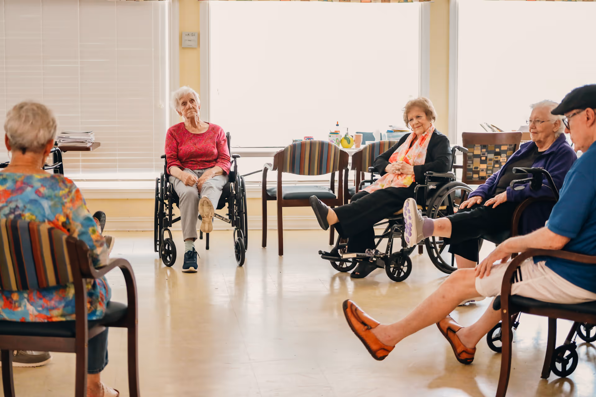 A group of elderly people, some in wheelchairs, sitting in a circle in a bright room with large windows, engaging in a seated exercise or stretching activity.