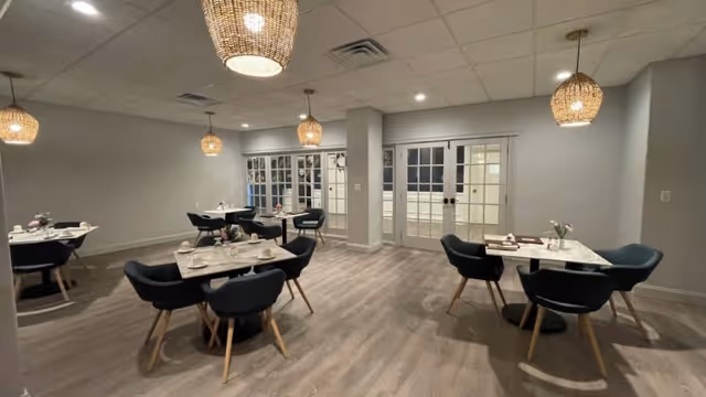A modern dining room with several small square tables, each surrounded by four dark upholstered chairs with wooden legs. The room has light wood flooring, gray walls, and multiple woven pendant light fixtures hanging from a white ceiling. In the background, there are glass double doors with white frames.