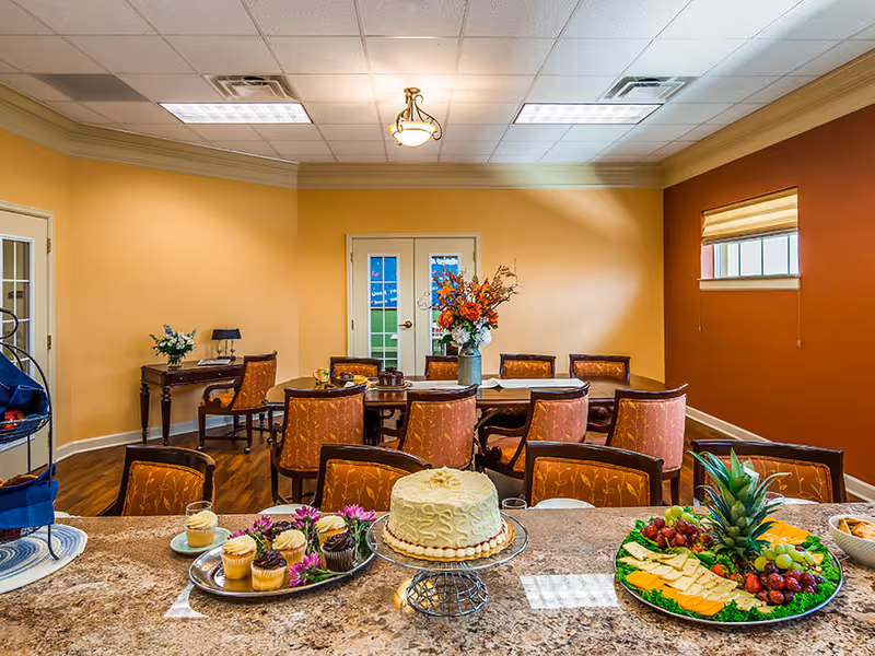 Communal dining room with a long table and chairs, a floral centerpiece, and desserts and fruit platters displayed on a foreground countertop.