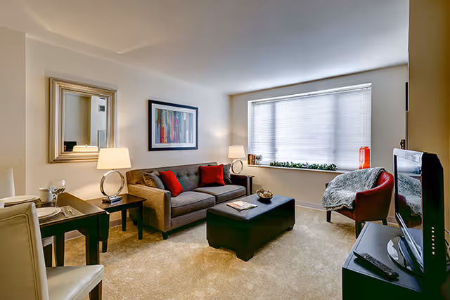 Well-lit living room with a gray sofa with red pillows, an ottoman coffee table, TV, armchair and a large window with blinds.