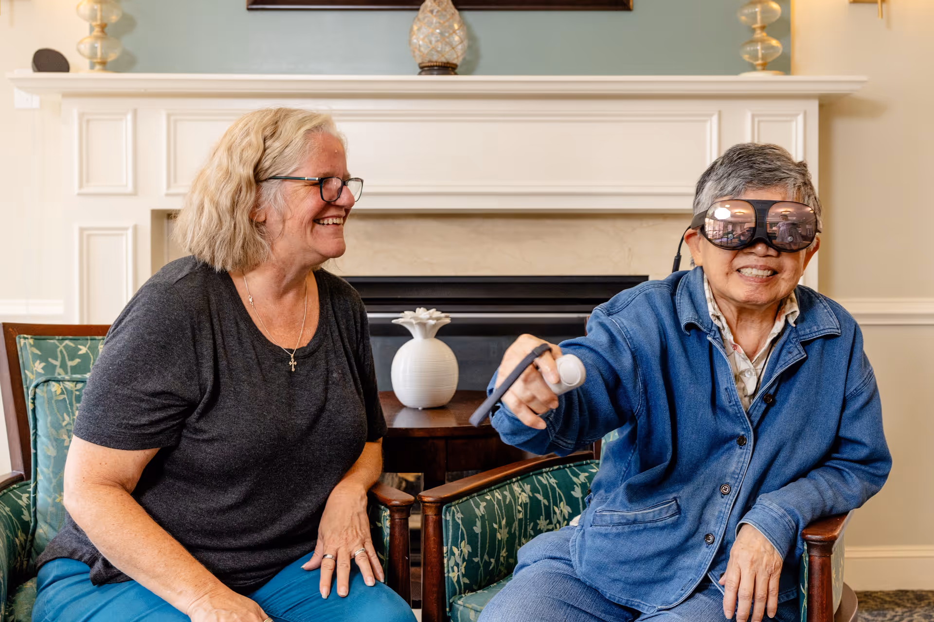 Two elderly women sitting in a living room area with a fireplace in the background. One woman is smiling and wearing glasses, while the other is wearing virtual reality goggles and holding a VR controller, also smiling.