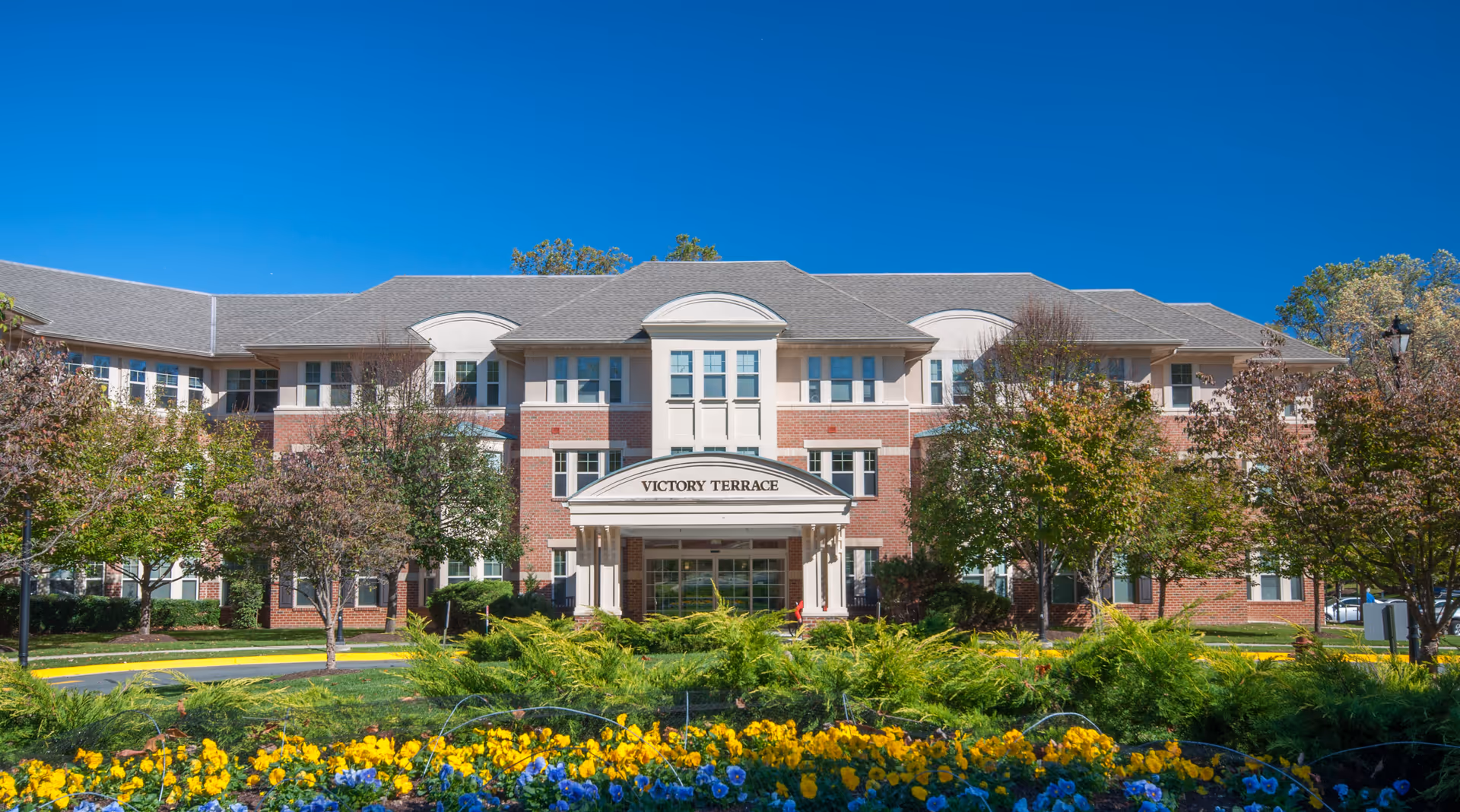 Front exterior view of Victory Terrace, a three-story senior living facility with a brick facade, white trim, and a covered entrance. The building is surrounded by trees and colorful flower beds under a clear blue sky.