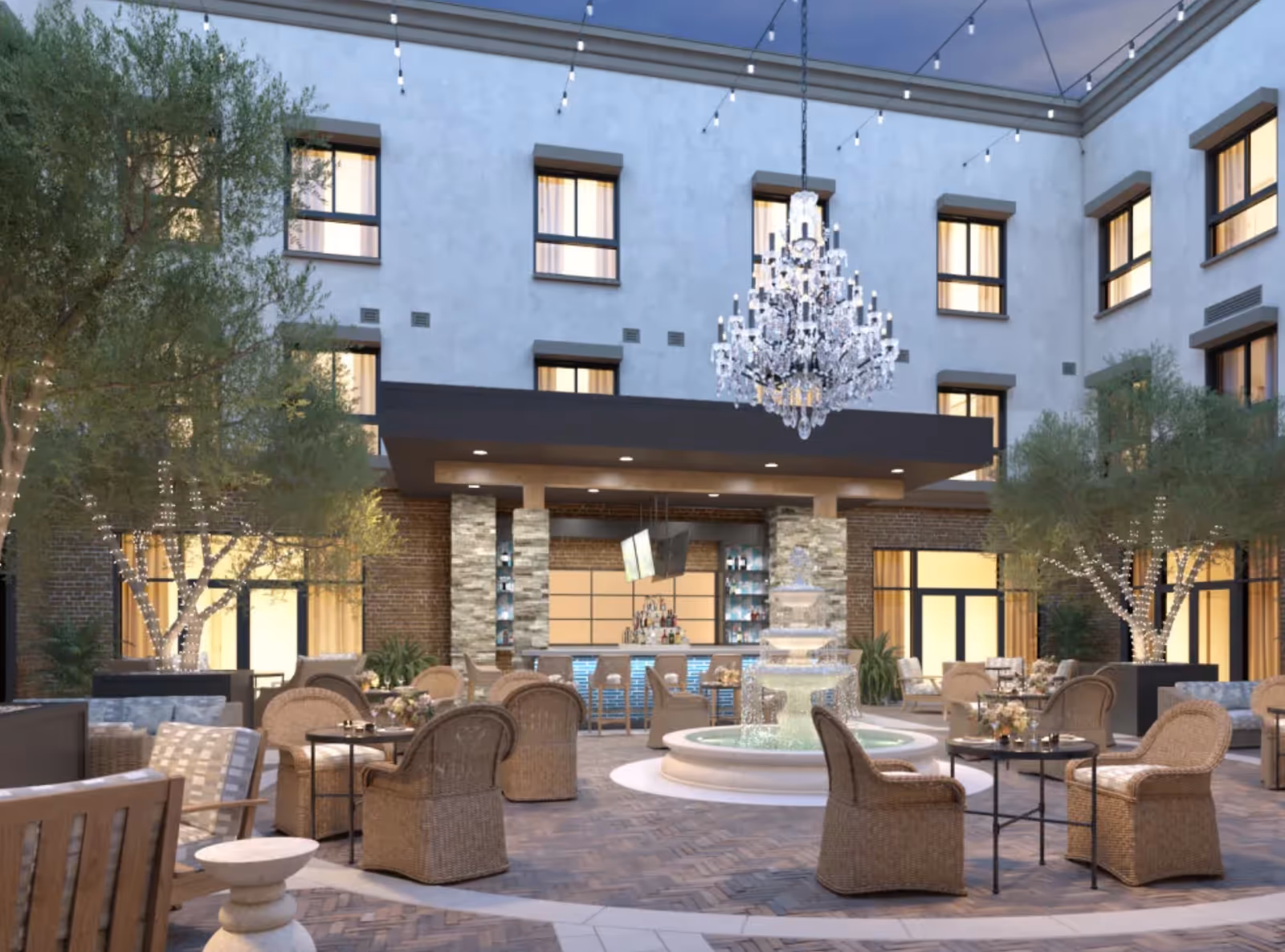 Outdoor courtyard area of Clearwater Newport Beach featuring wicker chairs and tables arranged around a central water fountain. The space is decorated with string lights and a large chandelier hanging above the fountain. Trees wrapped with lights and a bar area with stools are visible in the background, surrounded by a multi-story building with windows.