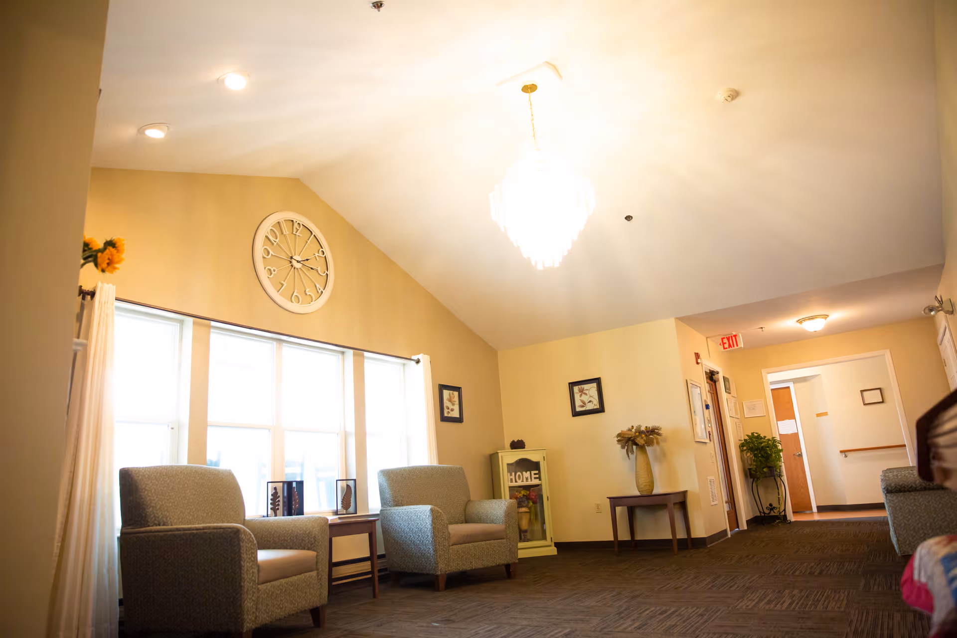 Bright senior living lounge with a vaulted ceiling and chandelier, two armchairs by large windows, a wall clock, and a hallway entrance.