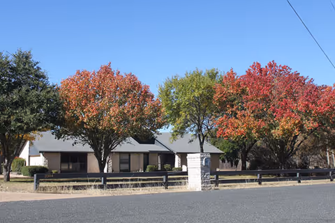 A single-story assisted living facility front with autumn trees and a low fence along the street.