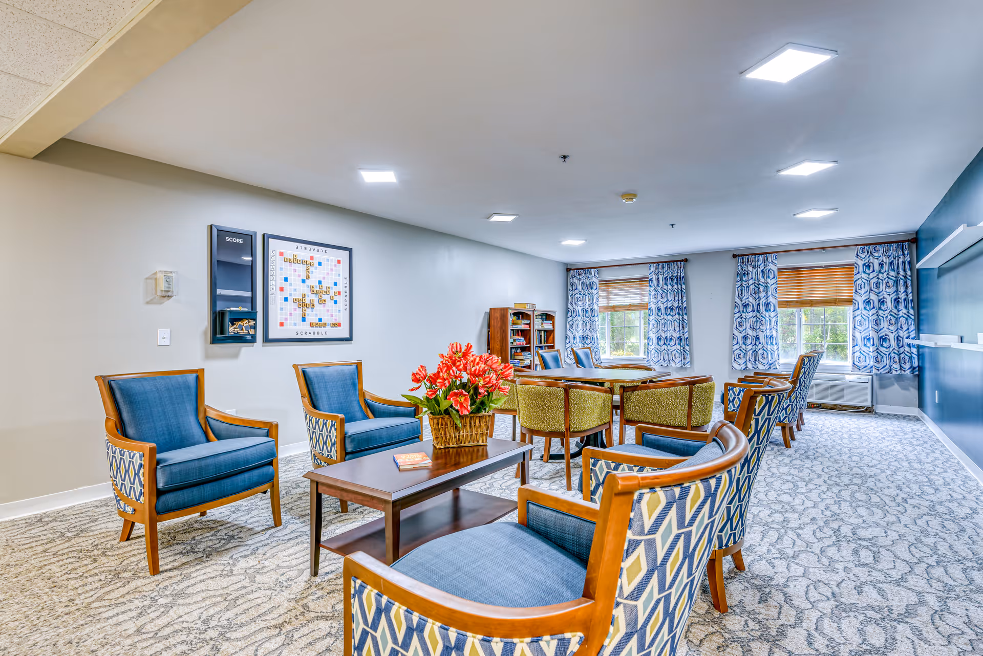 A bright and spacious common area in a senior living facility featuring multiple comfortable chairs with blue and patterned upholstery arranged around a wooden coffee table with a flower arrangement. In the background, there is a round table with additional chairs, bookshelves, and windows with patterned curtains allowing natural light to enter the room. A Scrabble game board is mounted on the wall.